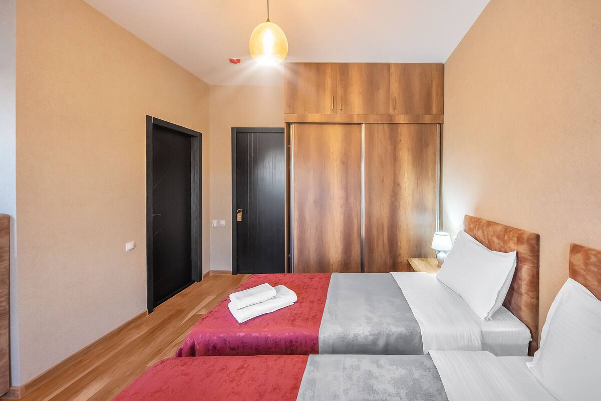 A hotel room with two beds, a wooden wardrobe, a nightstand with a lamp, and a hanging light fixture.