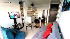 A living room with a blue sofa, a grey sofa with red cushions, a TV, a dining table with chairs, and a kitchen area in the background. - Confortable Apartamento Vacacional  (Ricaurte)