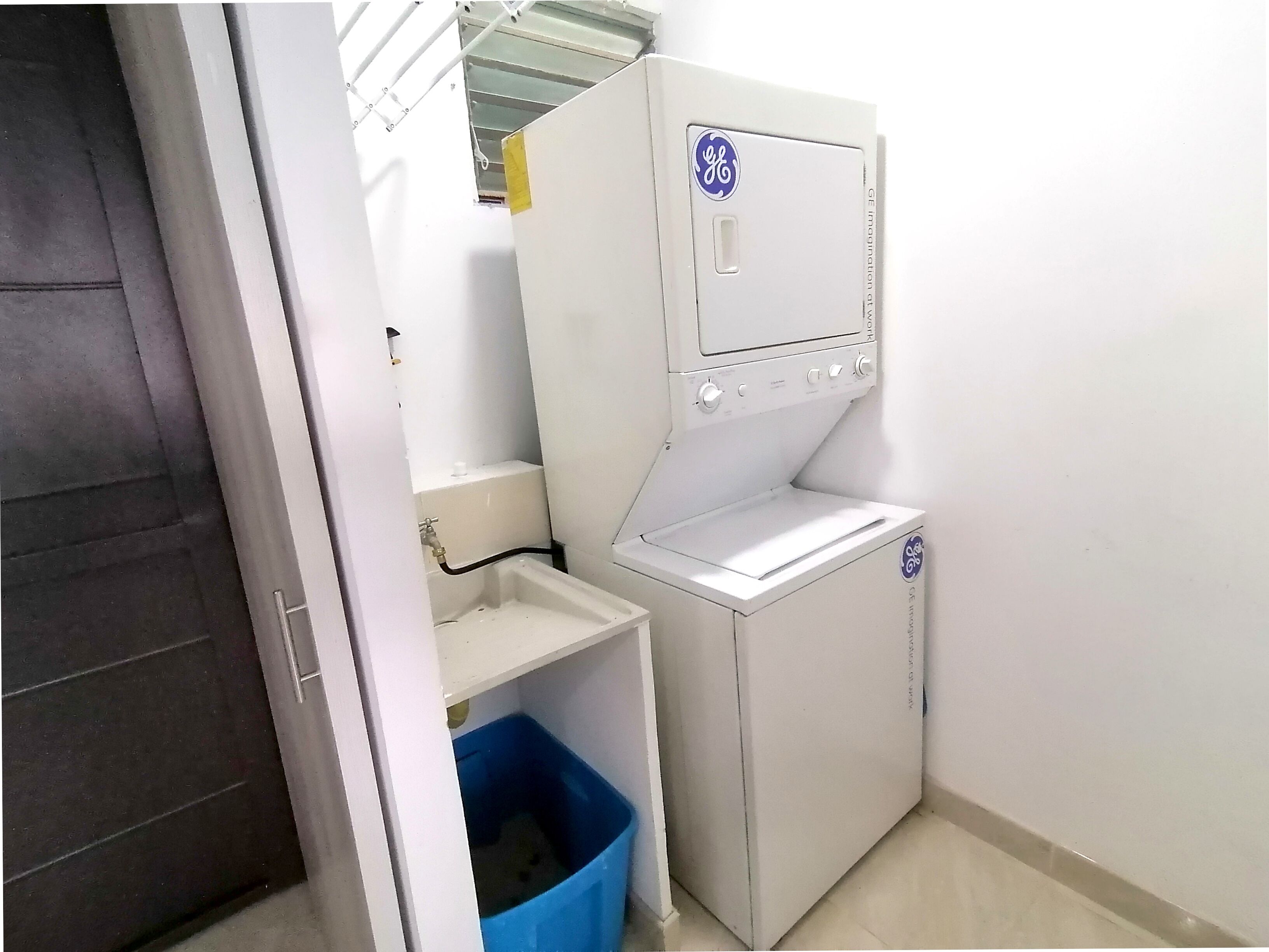 A compact, white laundry room with a GE brand washer and dryer, a small sink, and a blue trash bin.