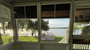 A room with a large window overlooking a lake, a wooden deck, and a grassy area. - Come and enjoy a family stay in a beatiful Lake House. (Lake Placid)
