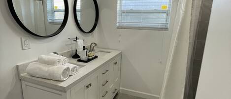 Un baño con tocador blanco, dos espejos, ducha con cortina blanca y ventana con persianas.