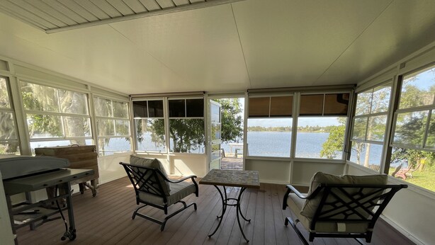 A covered porch with wooden flooring, two chairs, a table, and a bench. Large windows offer a view of a lake and trees. - Come and enjoy a family stay in a beatiful Lake House. (Lake Placid)