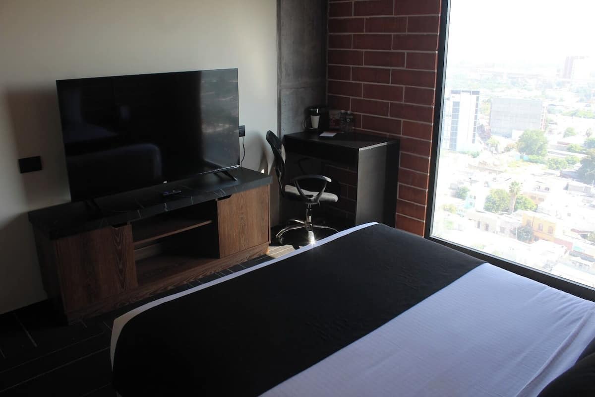 A hotel room with a bed, a TV on a wooden stand, a desk with a chair, and a window with a city view.
