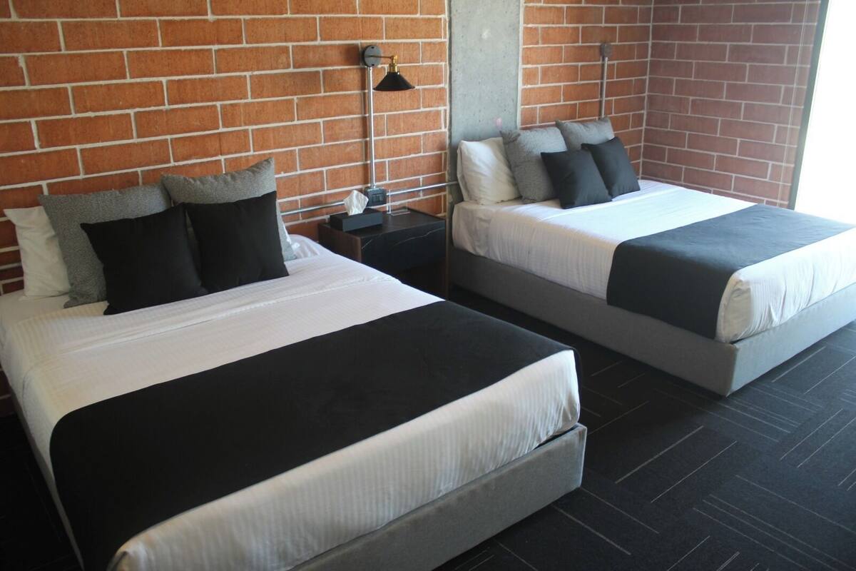 A hotel room with two beds, a brick wall, a modern lamp, and a concrete pillar.