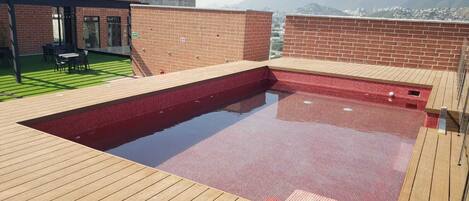 A rooftop swimming pool with a wooden deck, surrounded by brick walls and a view of distant mountains.