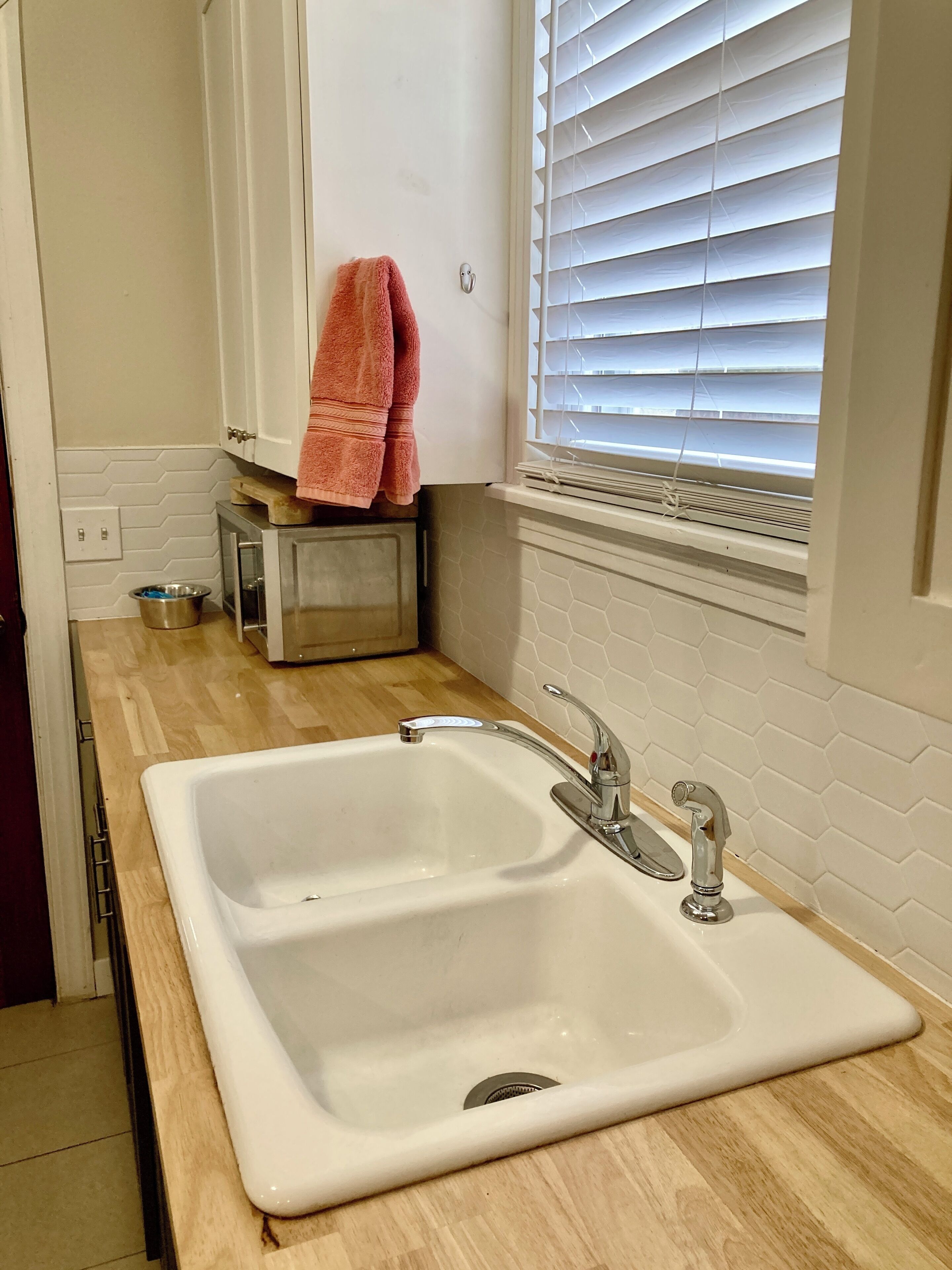 A kitchen with a white sink, two faucets, a window with blinds, a towel hanging on the cabinet, and a metal bowl on the countertop.
