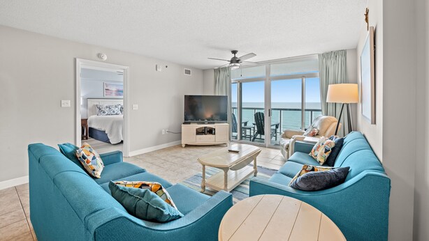 A spacious living room with a large TV, teal sofas, a coffee table, and a balcony with a view of the sea. - Oceanfront 3BD/3BA at Bluewater Keyes (806) (North Myrtle Beach)