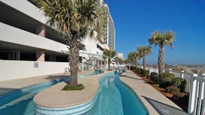 A swimming pool with a curved design, surrounded by palm trees and a white building with balconies. - Oceanfront 3BD/3BA at Bluewater Keyes (806) (North Myrtle Beach)