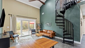 Un salon moderne avec une table à manger en bois, un canapé en cuir, un escalier en colimaçon noir et une grande fenêtre donnant sur l’extérieur.