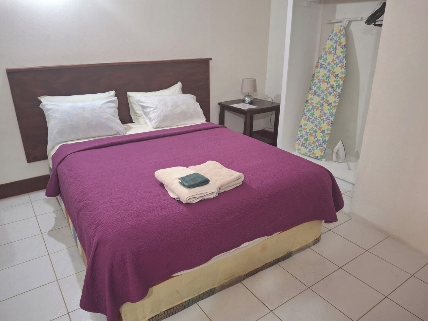 A neatly made bed with a purple bedspread and white pillows, a small bedside table with a lamp, and a shower curtain with a floral pattern.