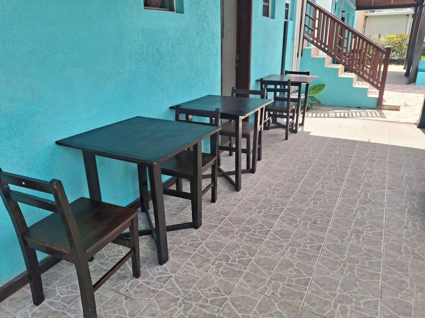 A row of dark wooden tables and chairs against a turquoise wall, with a tiled floor and a staircase in the background.