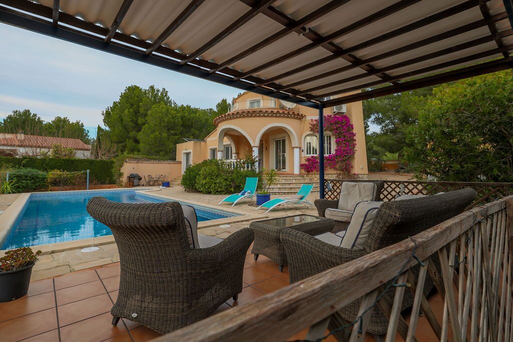 A covered outdoor area with a pool, wicker furniture, and a building with arches in the background.