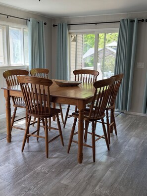 A dining area with a wooden table and chairs, large windows with curtains, and a view of greenery outside.
