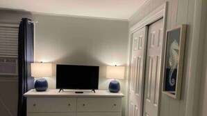 Een slaapkamer met een flatscreen televisie op een dressoir, twee lampen, een bed met een sprei, en een deur met decoratief paneel.