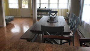 Een eetgedeelte met een houten tafel en stoelen, een keuken op de achtergrond, en een deur die naar een andere kamer leidt.