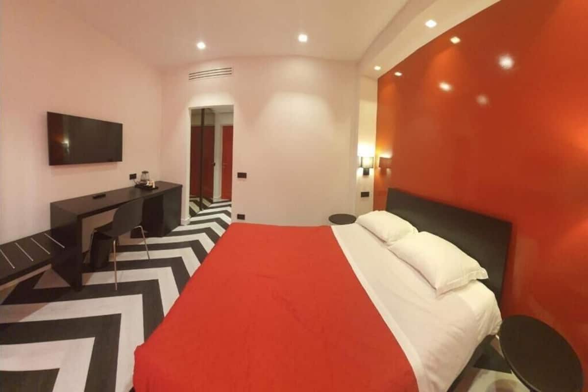A modern hotel room with a large bed, a desk, a flat-screen TV, and a red accent wall.