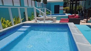 A rectangular pool with clear blue water, surrounded by a white and blue tiled deck. There are lounge chairs and a small staircase leading to a covered area. - BLR Beach Villas #12  (Double Bed) (Oistins)