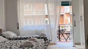 A hotel room with two beds, a large window with sheer curtains, a balcony with a table and chairs, and a hanging light fixture. - Monteverde Trastevere Dolce Vita Rooms (Roma)