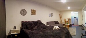 Un dormitori amb un llit, taules de nit, un escriptori i una cadira. Hi ha elements decoratius a la paret i una porta que dona a una altra habitació.