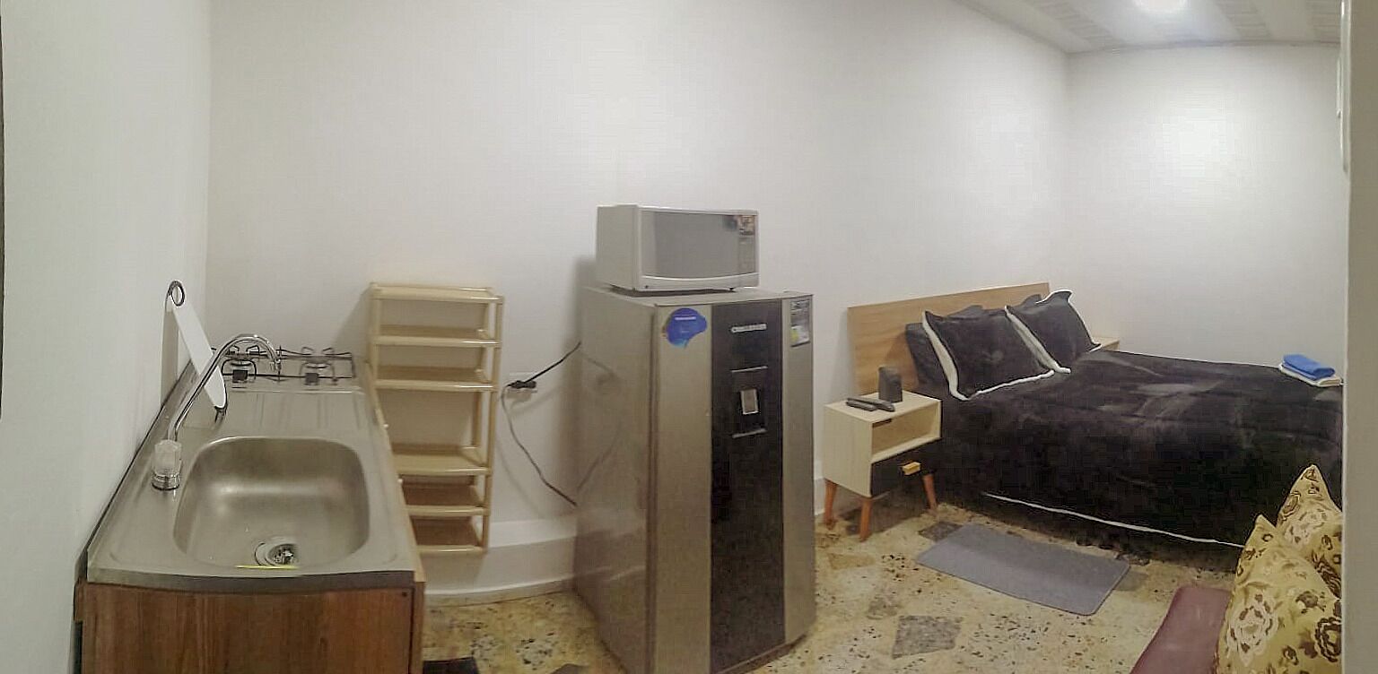 A compact hotel room with a kitchenette, a bed, a small bedside table, and a built-in microwave.