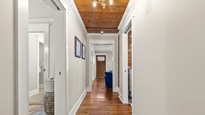 A hallway with wooden flooring, white walls, and a wooden ceiling. There are framed pictures on the walls and a blue chair in the distance. - Charming Historic Marietta Home! (Marietta)