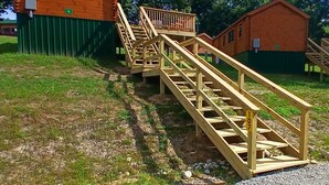 Eine Holztreppe führt zu einer Hütte mit einem grünen Zaun und blauem Himmel.