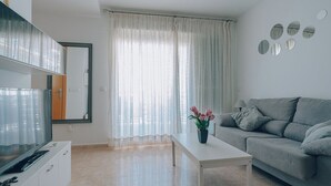 Una sala de estar moderna con una televisión de pantalla plana, un sofá gris, una mesa de centro blanca y un amplio ventanal con cortinas translúcidas.