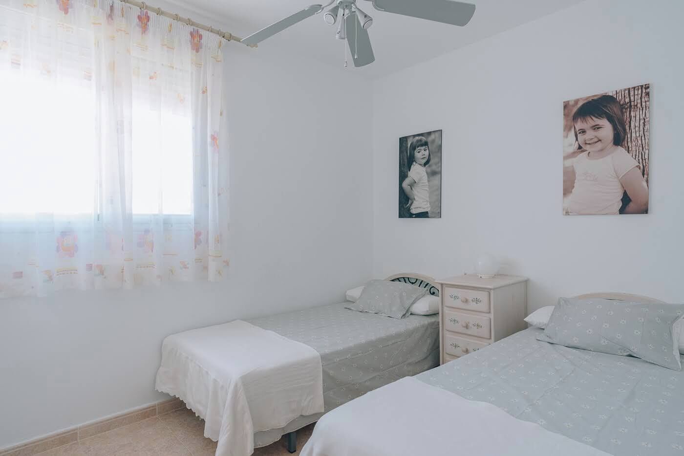 A bedroom with two single beds, a nightstand, a ceiling fan, and a window with curtains.