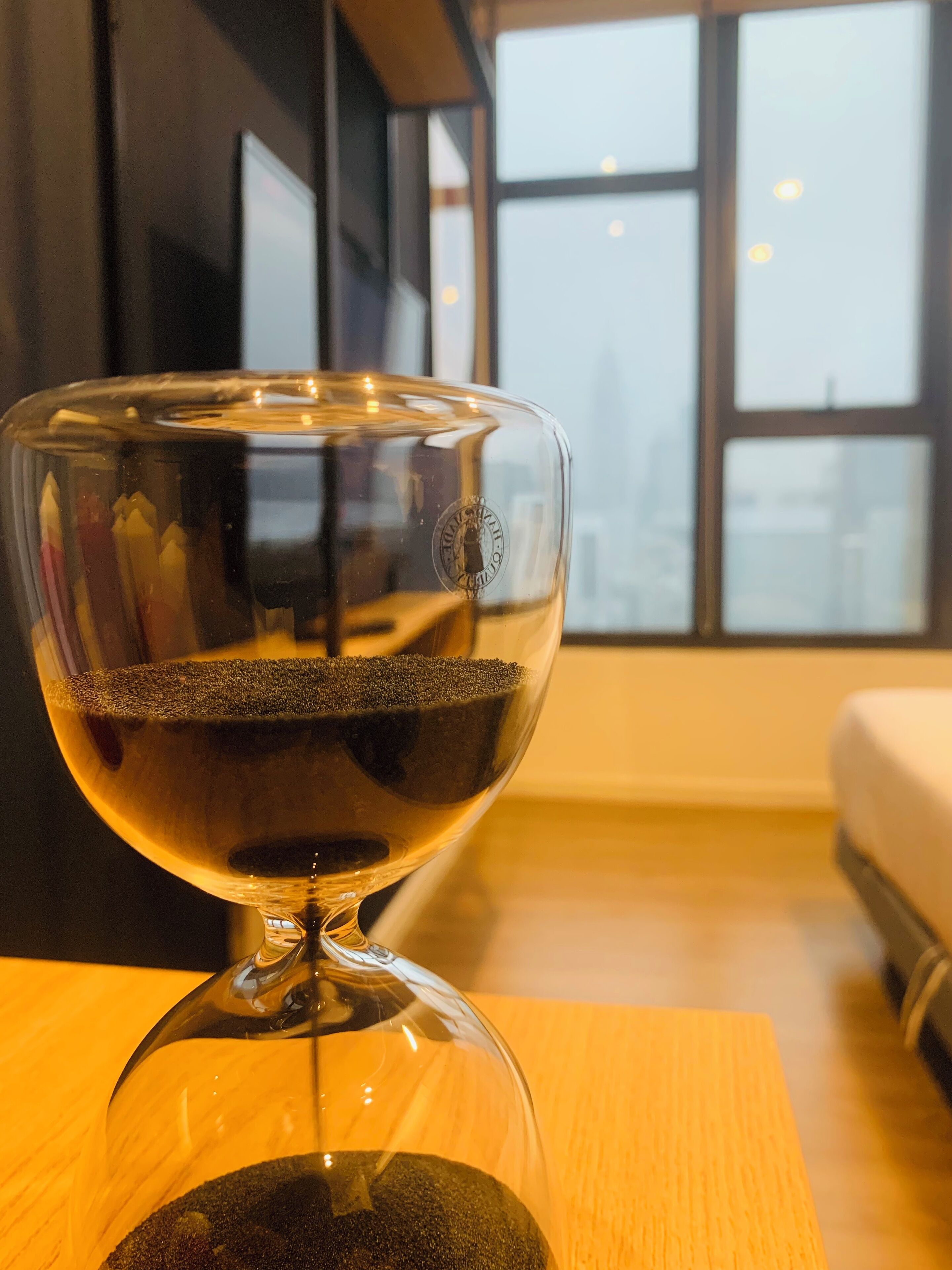A clear glass goblet with a dark liquid on a wooden table in a room with a large window and a bed in the background.