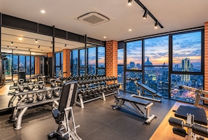 A gym with various exercise equipment, large windows with a city view, and a brick wall. - Ceylonz Suites by Roomy (Kuala Lumpur)