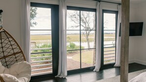 A room with a large window offering a view of a lake and trees. There is a wicker chair and a flat-screen TV mounted on the wall. - Stunning Marsh View Pool Retreat on St. Simons Island (St. Simons Island)