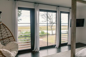 A room with a large window offering a view of a lake and trees. There is a wicker chair and a flat-screen TV mounted on the wall.