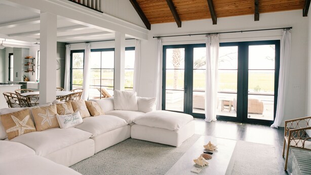 A spacious living area with a white sectional sofa, a dining table with chairs, and large windows with curtains. - Stunning Marsh View Pool Retreat on St. Simons Island (St. Simons Island)