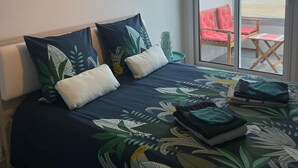 Un dormitorio con cama, almohadas, mesita de noche, ventana con vista al mar y una lámpara en el techo.