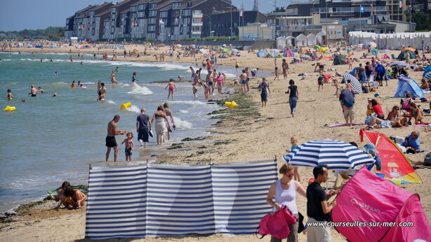 Una playa concurrida donde gente disfruta del agua y el sol. Se ven edificios al fondo y un cielo despejado.