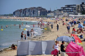 A crowded beach with people enjoying the water and sun. There are buildings in the background and a clear sky.
