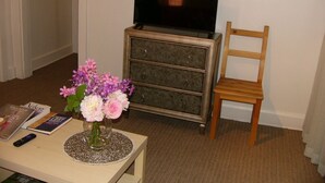 Ein Zimmer mit Fernseher auf einer Kommode, einem Holzsessel, einem Tisch mit Blumenvase und einigen Büchern.
