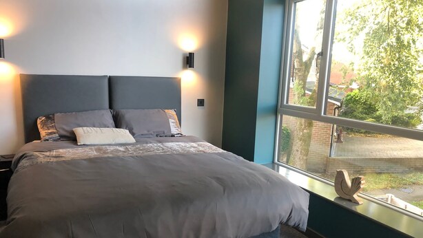 A bedroom with a large bed, two wall-mounted lights, a window with a view of the outside, and a decorative bird sculpture on the windowsill. - Welcome to Your Stay at The Nest (Bolton)