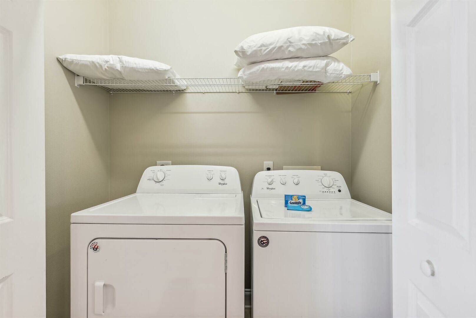 A laundry room with two white washers and dryers, a metal shelf above the dryers holding pillows, and a door to the left.