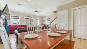 A dining area with a long wooden table set for four, white chairs, and four wine glasses. There is a ceiling fan, recessed lighting, and a red couch in the background.