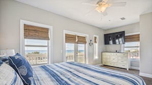 A bedroom with a large bed, a ceiling fan, a TV, and a view of the ocean.