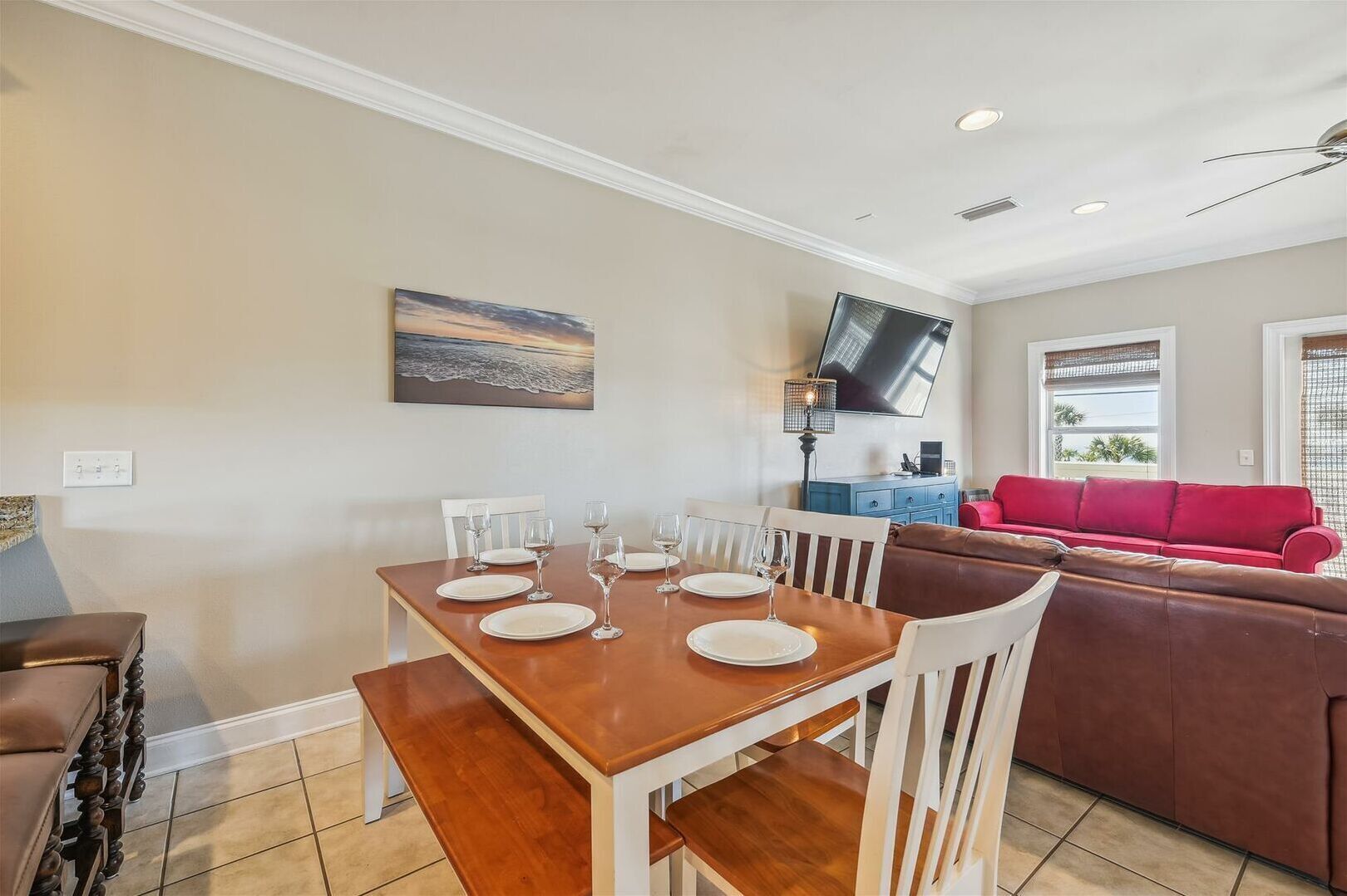 Un coin repas avec une table en bois dressée pour quatre personnes, des chaises blanches et un banc. Un canapé rouge et un meuble bleu sont visibles à l’arrière-plan.
