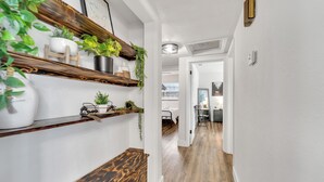 Um corredor com assoalho de madeira, paredes brancas e prateleiras com plantas. Há uma porta que dá para outro cômodo.