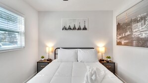 Cama bem arrumada com lençóis brancos, com duas mesas de cabeceira laterais, cada uma com luminária e pequenos vasos de plantas. Acima da cama, um quadro emoldurado com a Torre Eiffel.