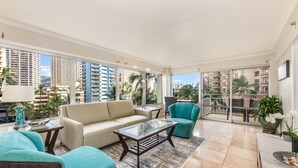 A spacious living area with a sofa, armchairs, a glass-top coffee table, and a view of tall buildings and palm trees through large windows. - Splendid 2BD w/ One FREE Parking (Honolulu)