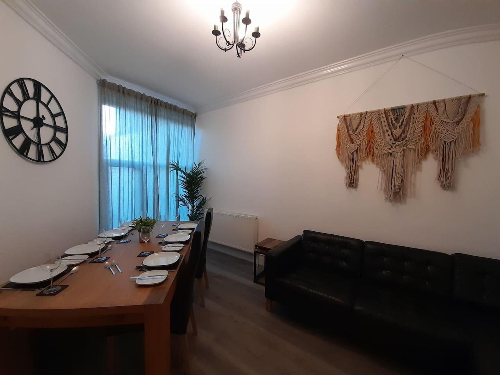 A dining area with a wooden table set for four, a black sofa, a large wall clock, and a macrame wall hanging.