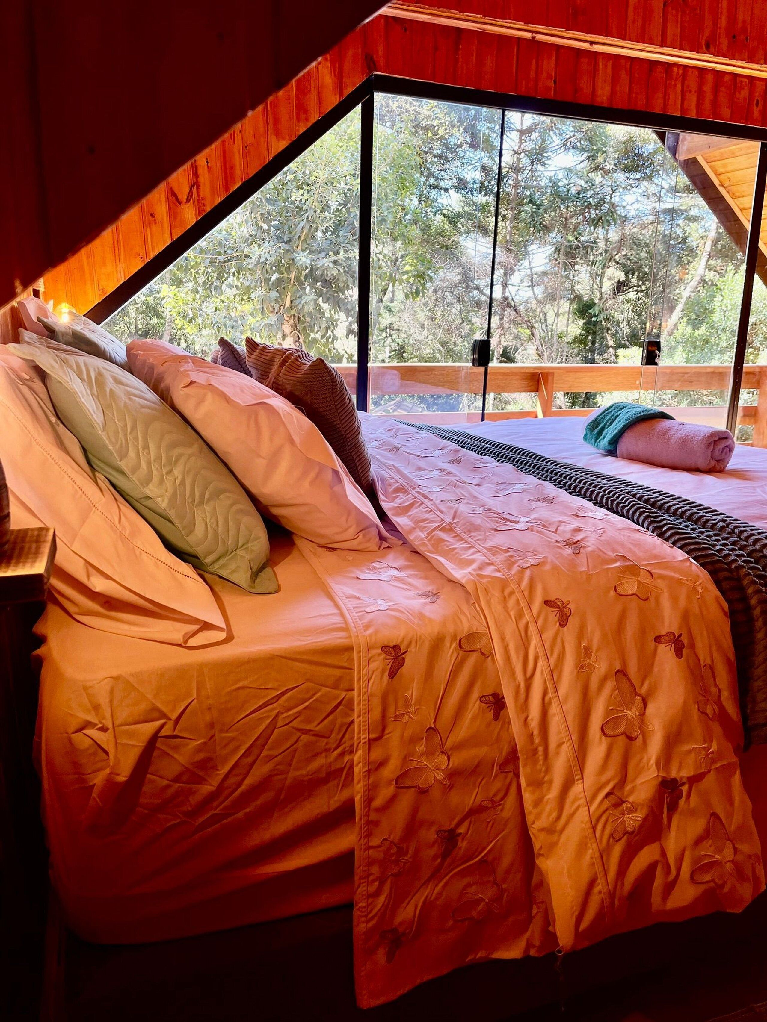 Un dormitorio con un ventanal grande, una cama con muchas almohadas y vistas a árboles.