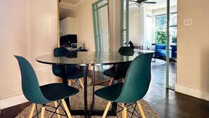 Un comedor moderno con una mesa de vidrio y sillas color turquesa, una cocina al fondo y vista a una sala de estar a través de un vano abierto.