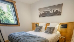 Ein Schlafzimmer mit einem großen Bett, einem hölzernen Kopfteil, einem an der Wand befestigten Fernseher und einem Fenster mit Blick auf Bäume.