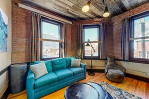 A room with a teal sofa, a round coffee table, and a leather armchair. There are large windows with curtains, a lamp, and a rug on the wooden floor. - Benson Lodge Room - 1 (Silverton)